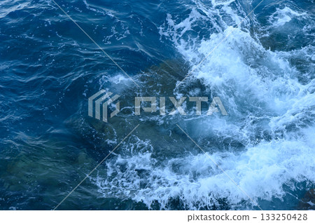 Waves crashing against rocky shorelines as sunlight dances on the ocean deep blue waters. 133250428