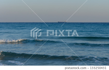 A tranquil ocean view with gentle waves and a distant cargo ship under a clear sky at sunset. 133250431