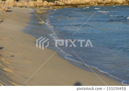 Tranquil shoreline at sunset with gentle waves lapping against golden sand and rocky cliffs. Tranquil shoreline at sunset with gentle waves lapping against golden sand and rocky cliffs. 133250458