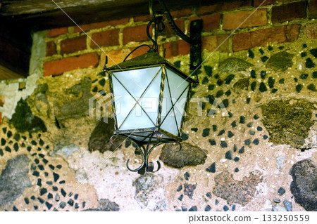 A forged retro lamp hangs on a stone wall, close-up, architecture A forged retro lamp hangs on a stone wall, close-up, architecture 133250559
