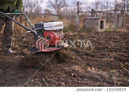 Adult using red motorized tiller for soil preparation in garden during fall 133250654