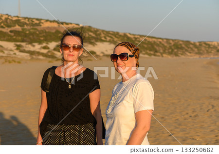 Two friends enjoying a sunset stroll along a serene beach in warm summer weather. 133250682
