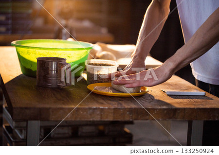 Hands kneading dough on yellow plate, preparing bread or pastry on wooden table with green bowl, metal sifter, and container of grains, warm light illuminating baking process Hands kneading dough on yellow plate, preparing bread or pastry on wooden table with green bowl, metal sifter, and container of grains, warm light illuminating baking process 133250924