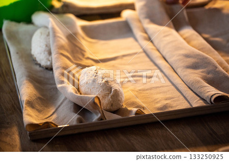 Raw dough resting on proofing cloth on metal tray during bread preparation process before baking, showcasing rustic texture of unbaked bread loaves Raw dough resting on proofing cloth on metal tray during bread preparation process before baking, showcasing rustic texture of unbaked bread loaves 133250925