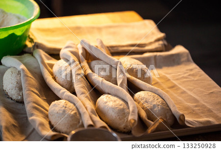 Raw bread dough rolls resting on linen cloth for proofing preparation of baking artisanal loaves organic flour seeds green bowl background warm light bakery kitchen setting 133250928