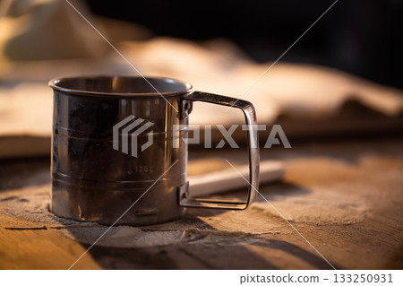 Metal flour sifter with measurement markings stands on wooden table, surface dusted with white flour, warm light illuminates baking tool for home cooking and confectionery preparation. 133250931