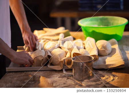 Baker hands preparing bread dough for baking process. Raw bread forms resting on floured linen cloths on wooden table. Rustic bakery scene with fresh dough and essential baking tools. 133250934