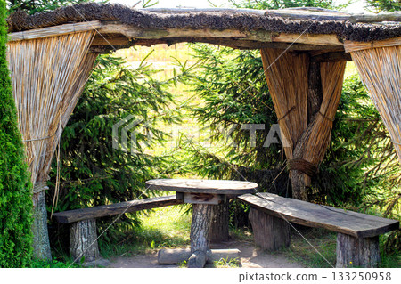 A beautiful gazebo made of dry reeds with wooden chairs and a table against the backdrop of fir trees in nature 133250958