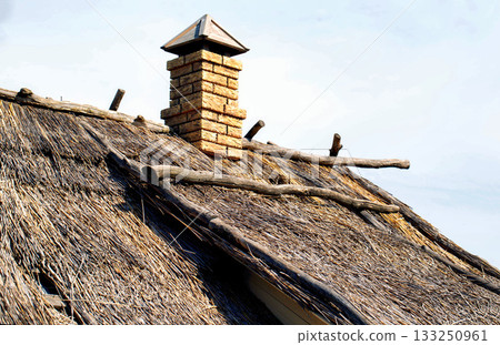 An old roof of a village hut with dry grass and reeds on the roof, a chimney against the sky, close-up. Copy space for text An old roof of a village hut with dry grass and reeds on the roof, a chimney against the sky, close-up. Copy space for text 133250961