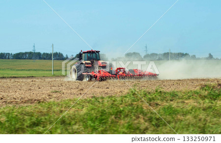 A modern tractor with a new cultivator cultivates the soil in a field before planting crops against a blue summer sky. Agricultural work on a collective farm. Copy space for text 133250971