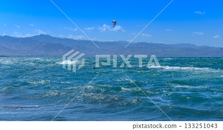 Kitesurfers ride the waves on a sunny day at the beach with mountains in the distance Kitesurfers ride the waves on a sunny day at the beach with mountains in the distance 133251043
