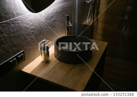 Stylish stone sink in a wooden countertop against the background of a mirror with diode lighting and a chrome tap. Fashionable interior, close-up 133251388