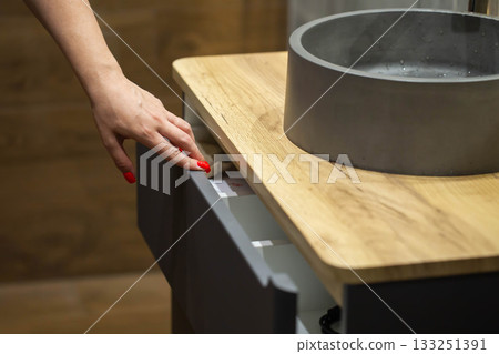 A woman's hand pulls out a shelf from a wall-mounted cabinet with a stone washbasin in a bathroom, close-up A woman's hand pulls out a shelf from a wall-mounted cabinet with a stone washbasin in a bathroom, close-up 133251391
