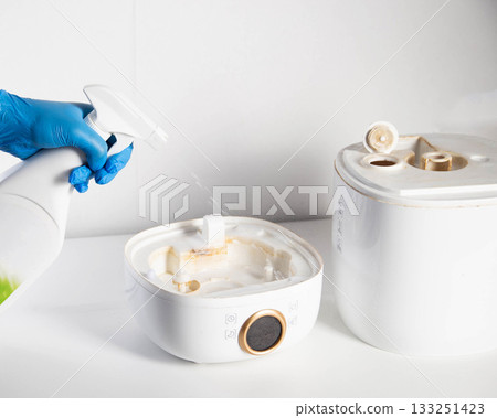Man in blue gloves spraying descaler, mold and odor remover into humidifier on white background. Humidifier device maintenance concept. Copy space for text 133251423
