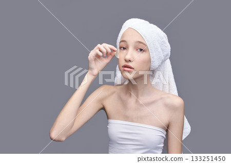 Young caucasian female applying skincare with cotton swab after shower on gray Young caucasian female applying skincare with cotton swab after shower on gray 133251450