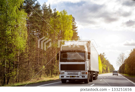 A modern truck transports cargo along a country road against the backdrop of beautiful nature and forest. Freight exchange. Transport and logistics company. Sunny weather. Copy space for text A modern truck transports cargo along a country road against the backdrop of beautiful nature and forest. Freight exchange. Transport and logistics company. Sunny weather. Copy space for text 133251656