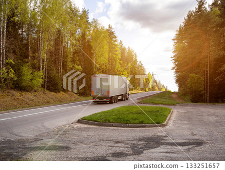 A truck with a semi-trailer transports cargo uphill in nature against the backdrop of a forest and the sun. Logistics company. Cost of cargo transportation. Cargo insurance. Copy space for text 133251657