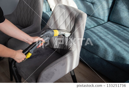 Cleaning a gray upholstered chair with a handheld steam cleaner in a modern living room with a blue couch Cleaning a gray upholstered chair with a handheld steam cleaner in a modern living room with a blue couch 133251736