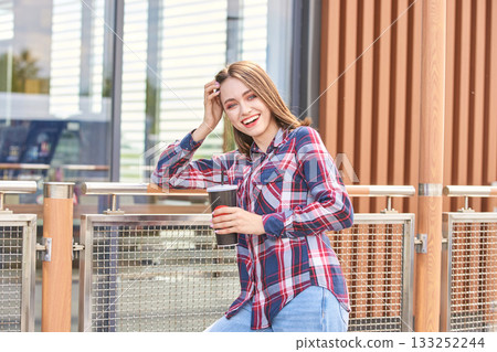 Smiling caucasian young female enjoying coffee on urban balcony in casual plaid 133252244