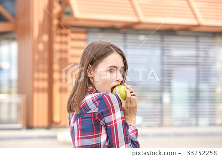 Caucasian young female eating green apple outdoors in casual plaid shirt 133252258
