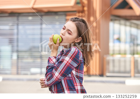 Young caucasian female enjoying green apple outdoors in casual plaid shirt 133252259