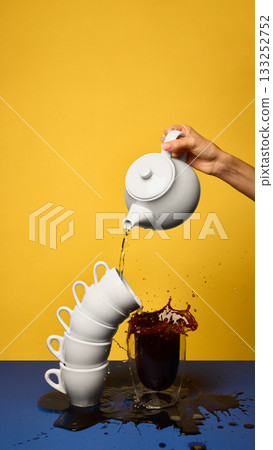 Stacked white cups tipping as coffee splashes into glass on yellow and blue setup 133252752