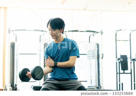 一位年輕男子正在健身房用啞鈴進行肌肉訓練。 133253669