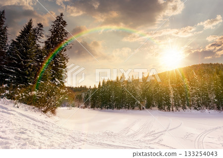 winter landscape with snow covered ground at sunset. beautiful mountain with frozen lake surrounded by spruce forest in evening light. wide view of winter wonderland. storytelling under the rainbow winter landscape with snow covered ground at sunset. beautiful mountain with frozen lake surrounded by spruce forest in evening light. wide view of winter wonderland. storytelling under the rainbow 133254043