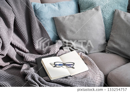 Open notebook with eyeglasses on cozy sofa with pillows and blanket 133254757