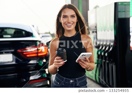 Fresh cup of coffee in hand, smartphone. A young woman at a gas station with her car 133254935
