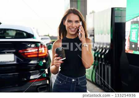 Fresh cup of coffee in hand, smartphone. A young woman at a gas station with her car Fresh cup of coffee in hand, smartphone. A young woman at a gas station with her car 133254936