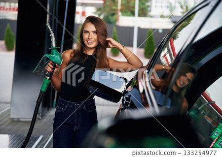 Leaning on the automobile, holding pump nozzle. A young woman at a gas station with her car 133254977