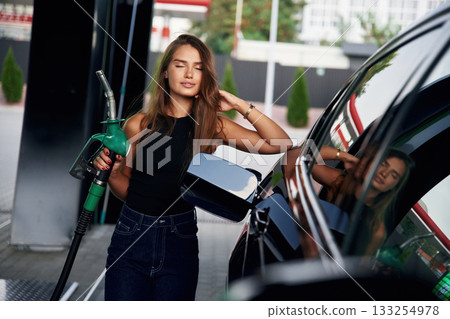 Leaning on the automobile, holding pump nozzle. A young woman at a gas station with her car 133254978