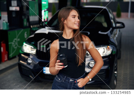 Black cup of the drink in hand. A young woman at a gas station with her car Black cup of the drink in hand. A young woman at a gas station with her car 133254985