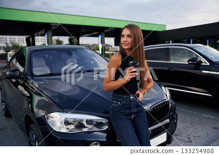 Front view, standing. A young woman at a gas station with her car 133254988