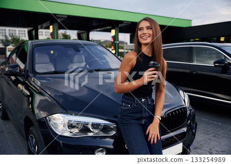 Front view, standing. A young woman at a gas station with her car Front view, standing. A young woman at a gas station with her car 133254989