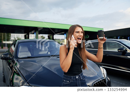 Happy conversation by phone. A young woman at a gas station with her car Happy conversation by phone. A young woman at a gas station with her car 133254994