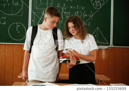 Girl and boy are standing, using smartphone. Pupils, kids in the classroom together Girl and boy are standing, using smartphone. Pupils, kids in the classroom together 133255609