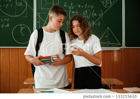 Girl and boy are standing, using smartphone. Pupils, kids in the classroom together 133255610
