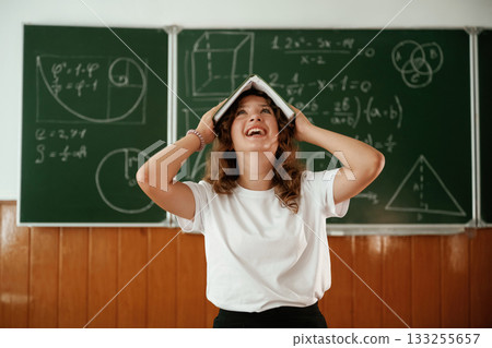 Fun, with notepad over the head. Schoolgirl is standing against blackboard in the classroom Fun, with notepad over the head. Schoolgirl is standing against blackboard in the classroom 133255657