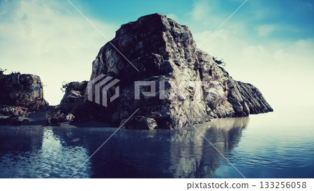 In a tranquil setting, a large rock formation rises from the calm water, reflecting the bright blue sky and soft clouds. The coastal scene evokes a sense of peace and natural beauty. 133256058