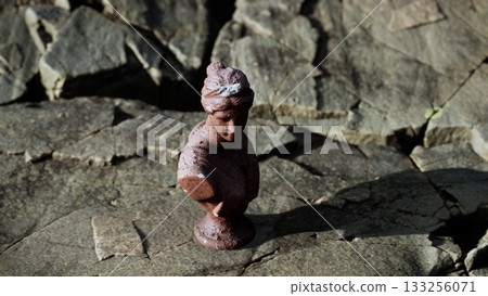 A small brown sculpture of a woman stands on a rough, rocky surface. Sunlight highlights the details while shadows create a striking contrast. This scene captures a moment of quiet beauty outdoors. 133256071