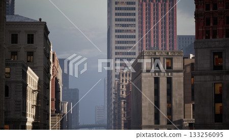 Tall buildings stretch towards the sky as the sun sets, casting a warm glow over the urban landscape. The streets below are quiet, creating a serene atmosphere in the city. 133256095