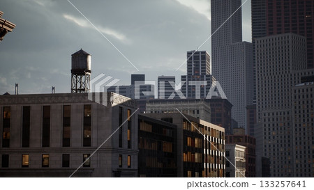 Dusk settles over an urban landscape, highlighting a blend of modern skyscrapers and a vintage water tower amid the city. Shadows stretch over the buildings as lights begin to glow. Dusk settles over an urban landscape, highlighting a blend of modern skyscrapers and a vintage water tower amid the city. Shadows stretch over the buildings as lights begin to glow. 133257641
