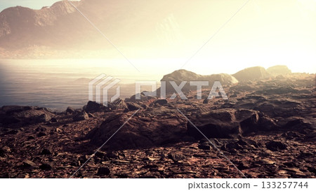 Soft light bathes a rugged shoreline as gentle waves lap against the rocks. Mist envelops distant mountains, creating a peaceful atmosphere at dawn. Nature’s beauty invites contemplation. 133257744