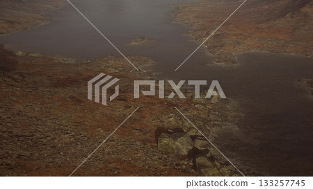 Rippling waters calmly meet rocky shores in a tranquil landscape during twilight. The warm hues of dawn reflect softly on the surface, creating a peaceful atmosphere. 133257745