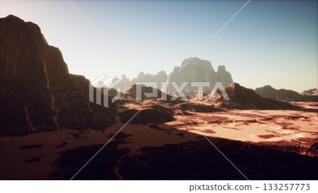 Under a bright blue sky, a wide desert spreads out with majestic mountains rising in the distance. The sun casts long shadows on the rocky terrain, highlighting the beauty of the natural scenery. 133257773