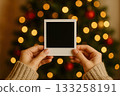Hands holding a blank photo in front of a decorated Christmas tree with lights 133258191