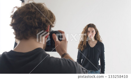 Behind the Scenes: Photographer with DSLR Camera Shoots a Serious Young Female Model with Long Curly Hair Against a Clean White Backdrop in a Professional Studio Photoshoot Behind the Scenes: Photographer with DSLR Camera Shoots a Serious Young Female Model with Long Curly Hair Against a Clean White Backdrop in a Professional Studio Photoshoot 133258410