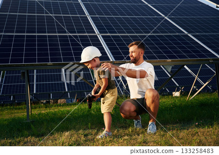 In hard hat and with drill. Father with his little son are together near the solar panels In hard hat and with drill. Father with his little son are together near the solar panels 133258483